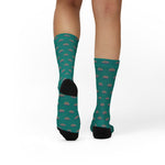 Rear view of a model's calves in teal Quahog Squad funny beach socks, black heel cups against the clam shell print