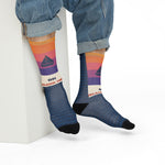 Model in jeans seated on a white block showing the sailboat and On Island Time cuff on Block Island socks