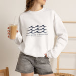 Woman holding iced coffee in a white One More Crossing Block Island sweatshirt and denim shorts