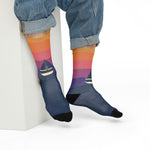 Seated model in rolled jeans leaning on a white block in orange-to-navy sunset sailboat socks