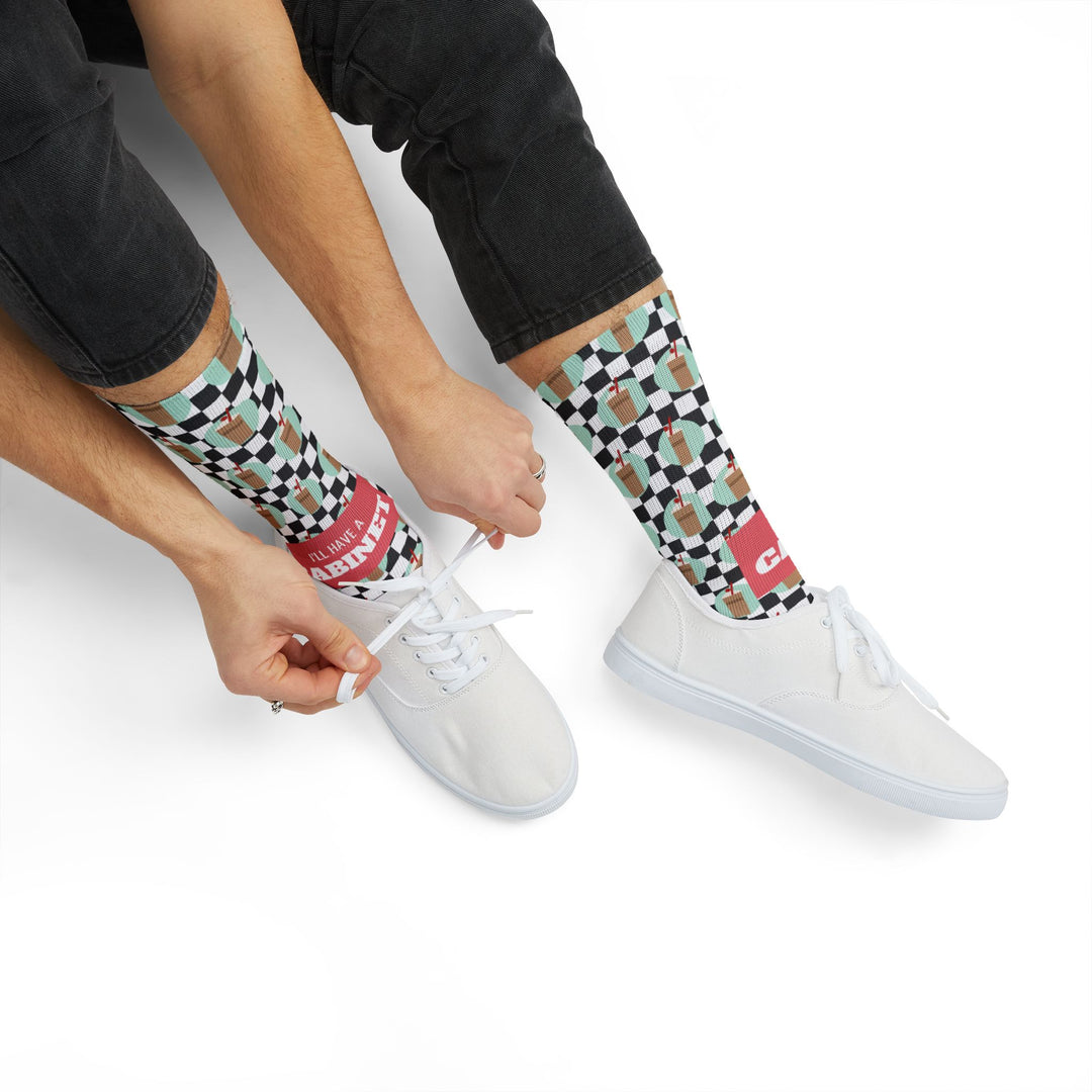 Seated model in black jeans tying white sneakers over I'll Have a Cabinet Rhode Island socks, coral band at the ankle