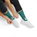 Seated model in black jeans tying white sneakers over teal Quahog Squad Rhode Island socks, cream band at the ankle