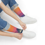 Seated model tying white sneakers while wearing sunset fade nautical crew socks with sailboat graphic