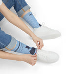 Seated model in rolled jeans pulling on light blue Block Island socks next to white sneakers