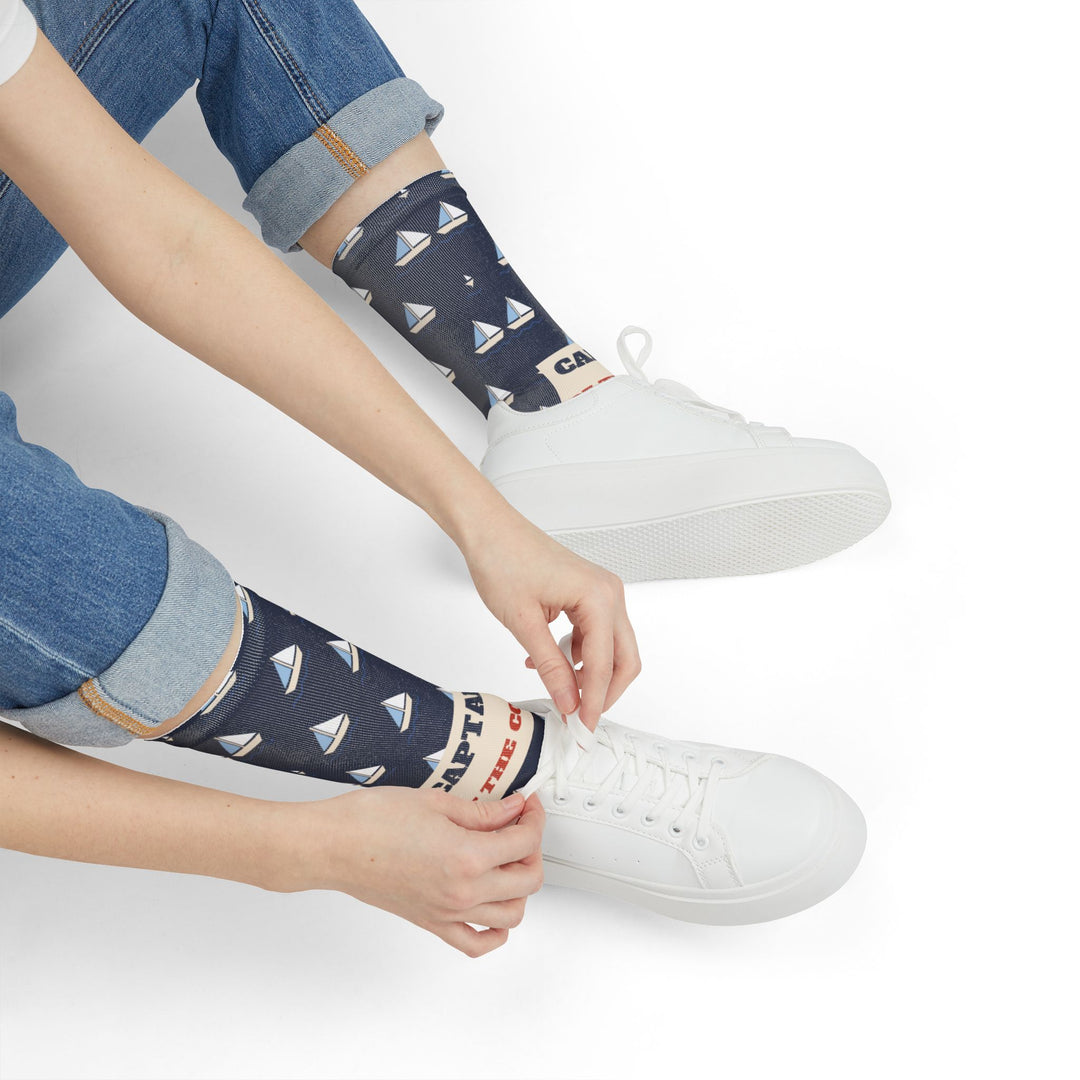 Seated model tying a white sneaker while wearing navy Captain of the Couch funny sailboat socks