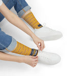 Seated model in rolled jeans pulling on I Like My Wieners All the Way funny beach socks beside sneakers